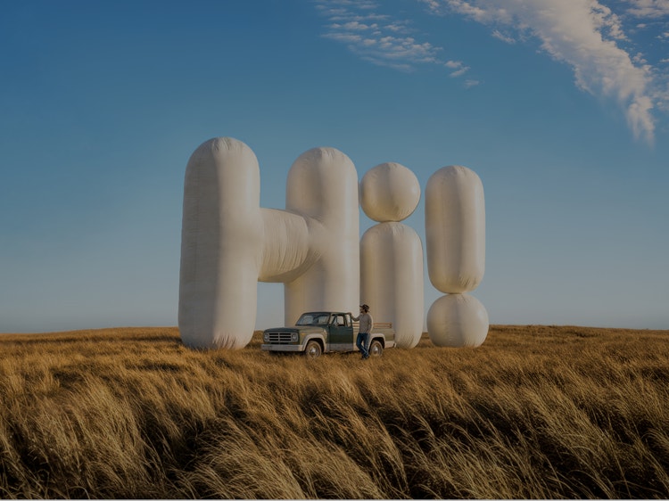 Large inflatable-style letters spelling &#39;Hi!&#39; standing in a wheat field with farmer and truck standing in front