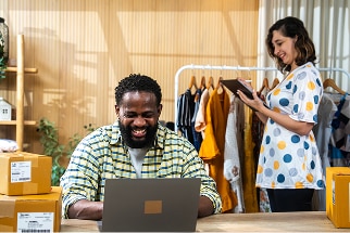 Man typing on laptop with a woman walking by behind him, looking down at a tablet