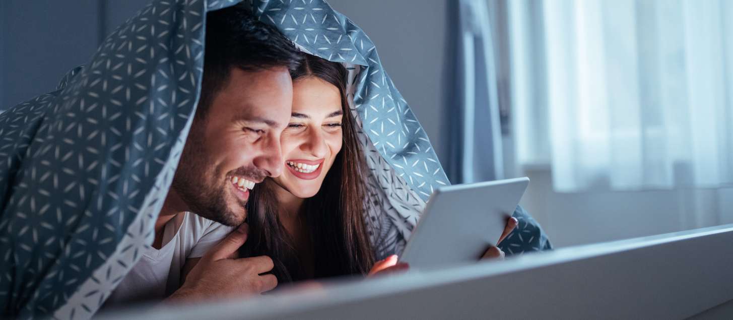 Man and woman looking at a tablet with a blanket over their heads.