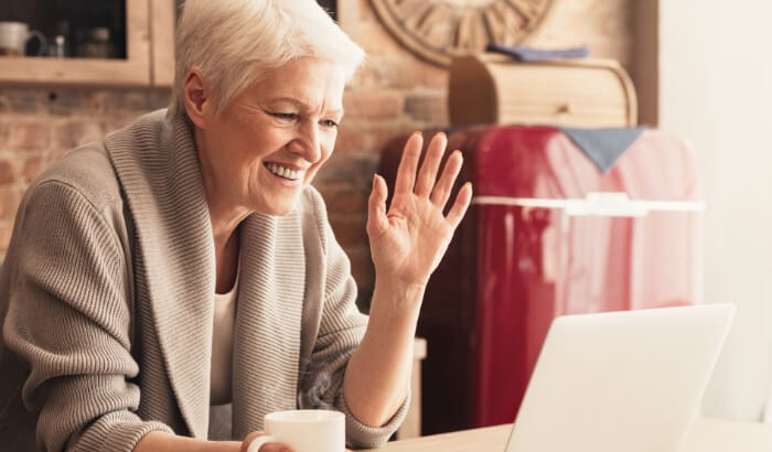 An older woman sitting at a table, drinking a coffee, video-calling on her laptop while connected to Spectrum Internet.