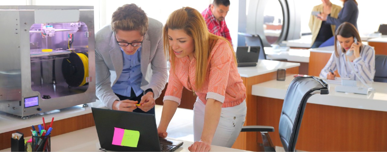 Two business people working on a laptop in an office with other coworkers working at their desks in the background.