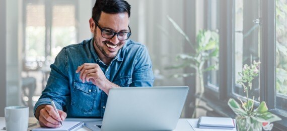 A smiling man wearing glasses sits at a desk by a window, looking at a laptop screen while holding a pen over a notebook.
