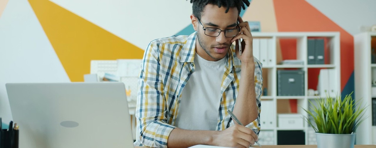 Male employee is speaking on mobile phone in office writing in notebook and smiling discussing business at work. 