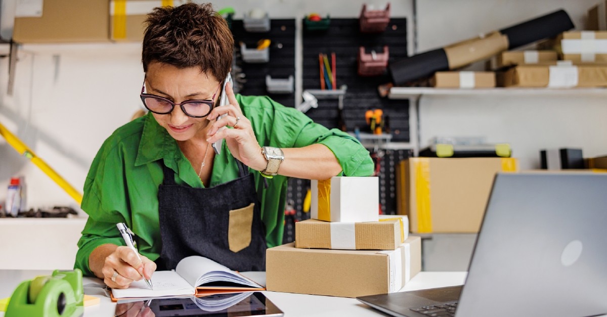 Male employee is speaking on mobile phone in office writing in notebook and smiling discussing business at work. 