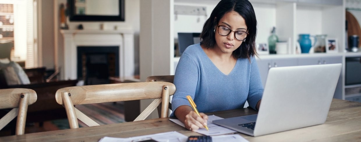Business woman works at kitchen table on laptop, while taking notes on a notepad