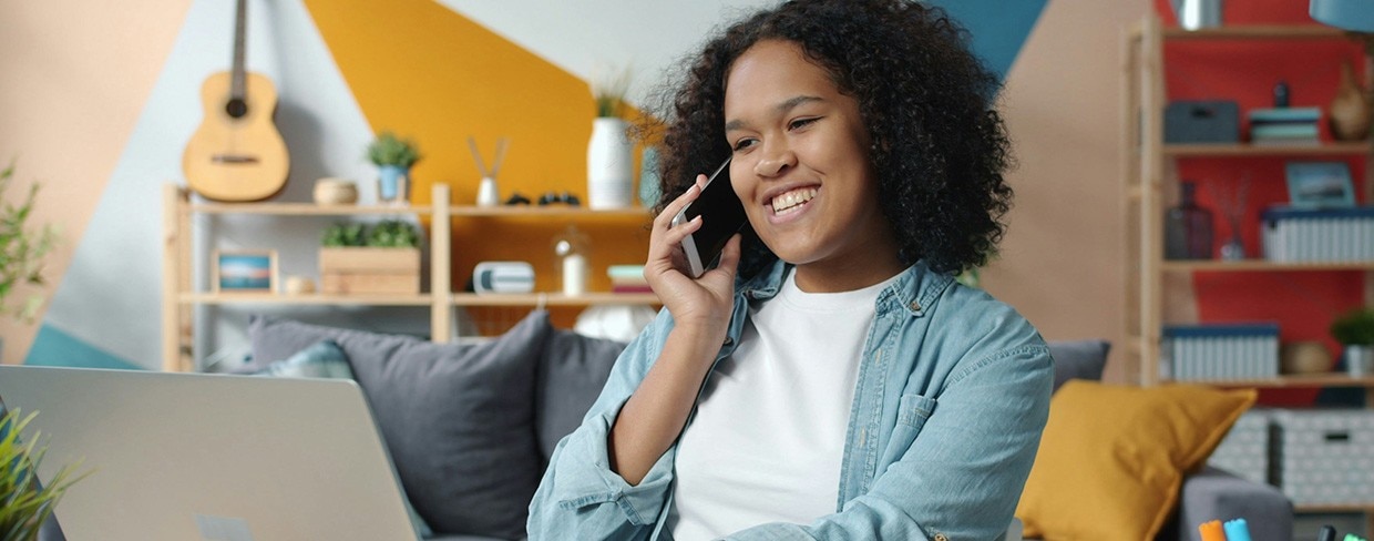 Young woman talking on mobile phone while smiling and working with laptop at home