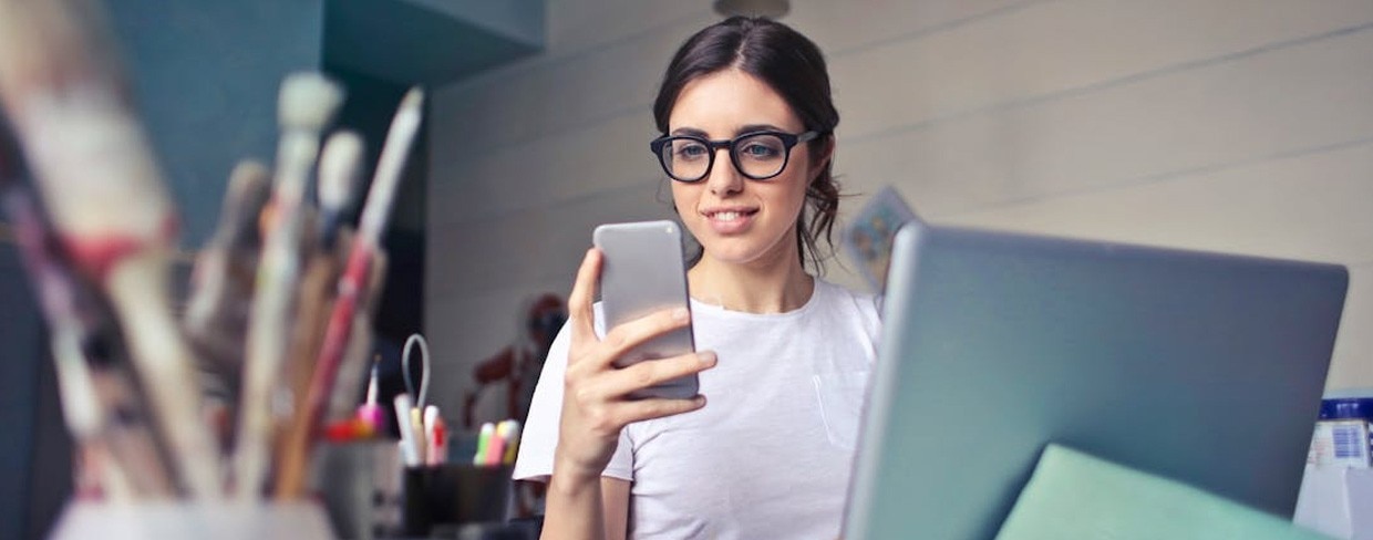 Woman wearing glasses smiling while looking at her smartphone, sitting at a desk with a laptop and art supplies nearby.