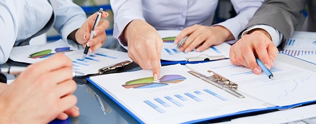 Several business people working together at a desk, pointing at charts in a binder in front of them.