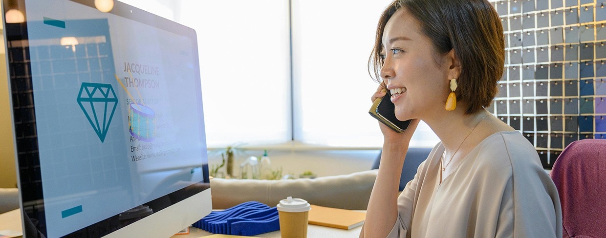 Smiling woman talking on a smartphone while working at her computer in a bright modern office, representing business communication and productivity.