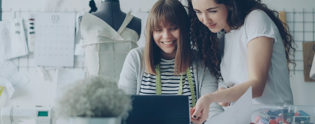 Female employee of tailor shop is measuring garment drawings with sewing threads while her attractive colleague is showing her tablet screen and talking to her.