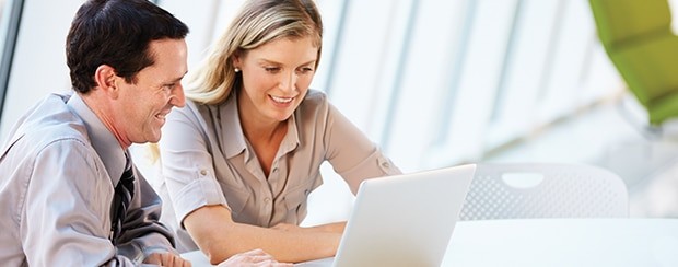 A business man and business woman smile and talk while working on a laptop.