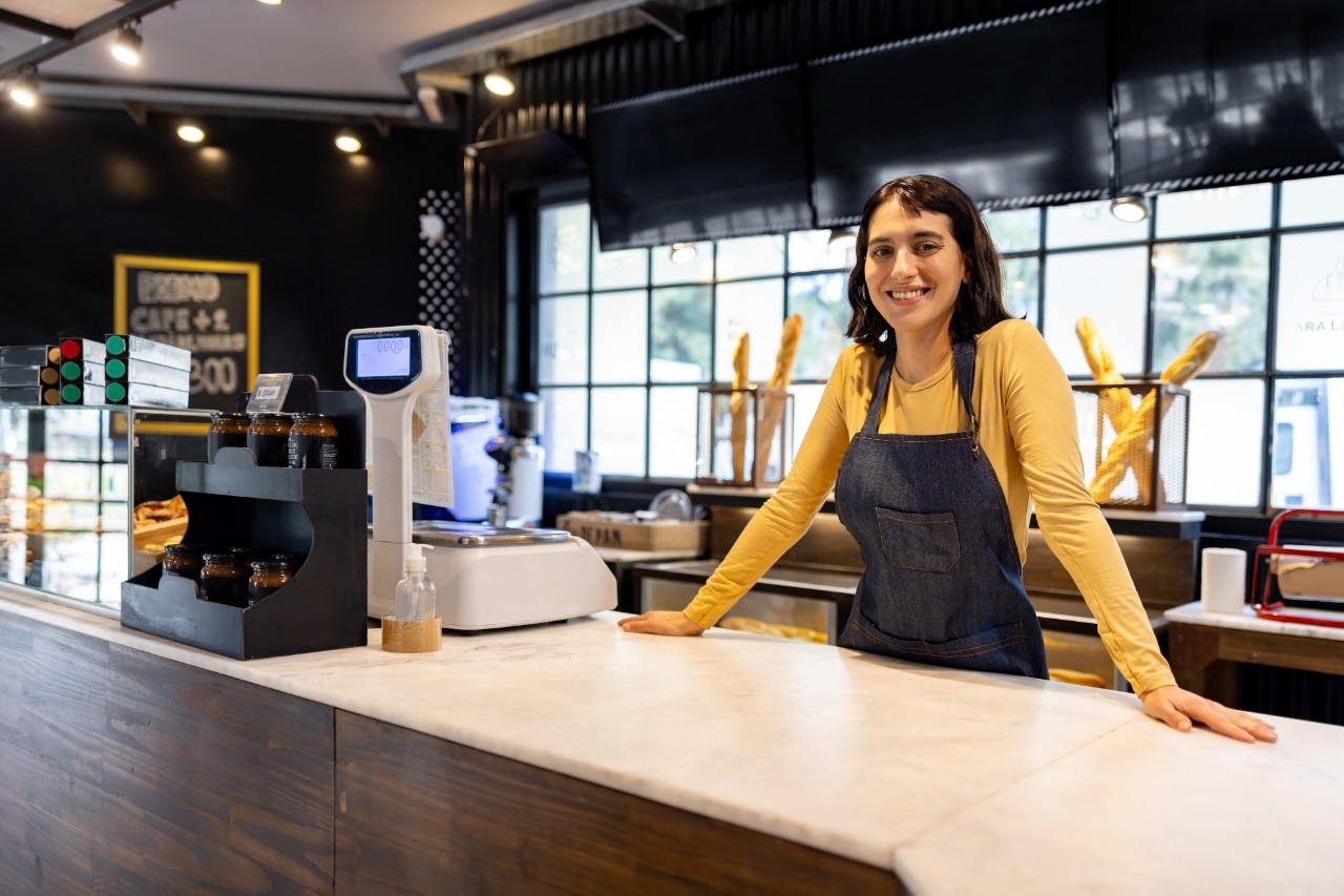 Happy retail clerk working at a bakery and smiling behind the counter while looking at the camera - small business concepts