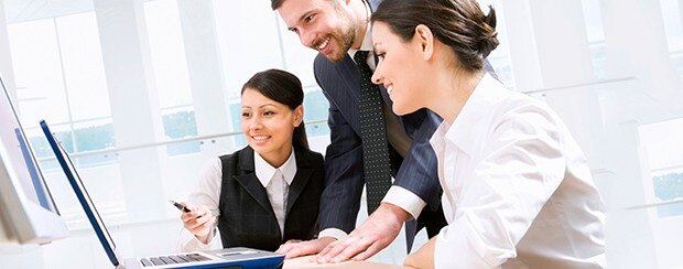A businessman in a suit uses a laptop, while talking with two female coworkers on either side of him.