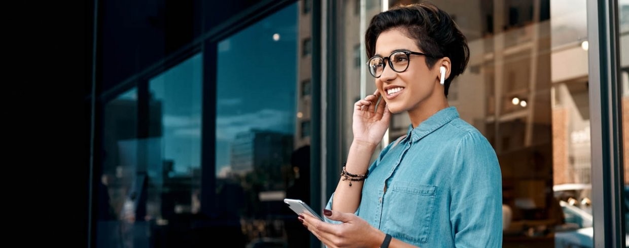 Smiling young woman wearing wireless earbuds and glasses, holding a smartphone, talking outside a modern glass building.
