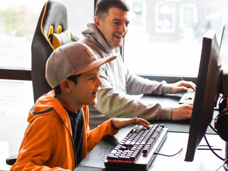 Un niño con una sudadera naranja con capucha y una gorra sonríe ante un teclado mientras un hombre se ríe frente a una computadora detrás de él.