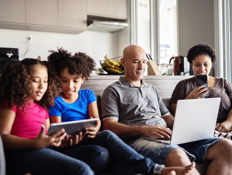 Una familia de cuatro se sienta junta en un sillón con una laptop y tabletas.