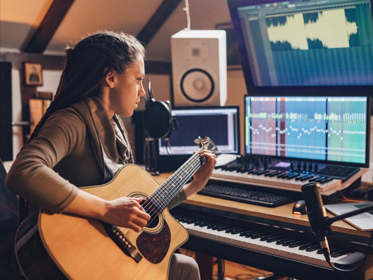 Una mujer toca una guitarra acústica en un estudio de música donde hay un teclado y pantallas de computadora.
