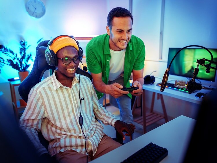 Un hombre sonriente con un gorro naranja está sentado frente a la computadora mientras un amigo está de pie detrás jugando videojuegos.