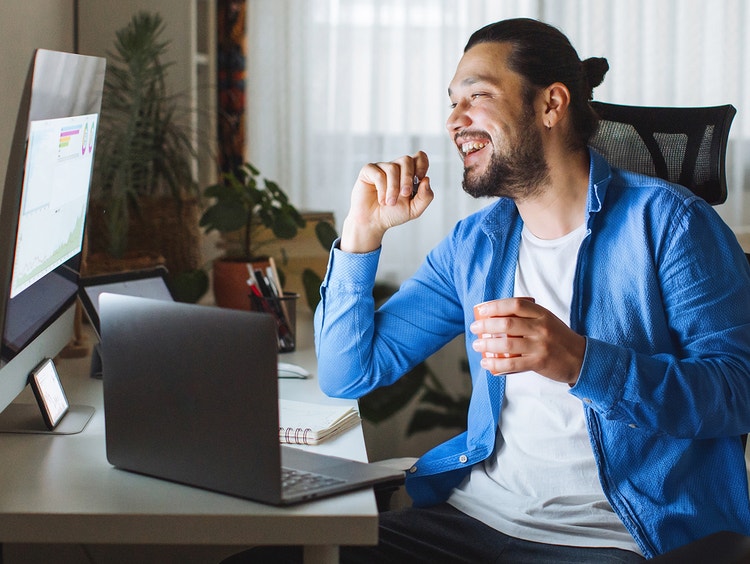 Un hombre sonriente con una chaqueta azul está sentado frente a un escritorio con una laptop y un monitor.