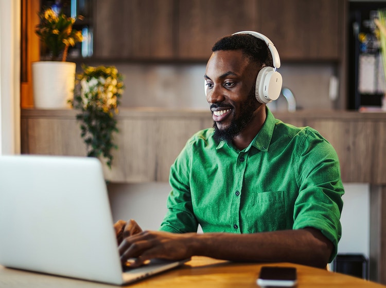 Un hombre sonriente con auriculares trabaja en una laptop en una mesa de la cocina.