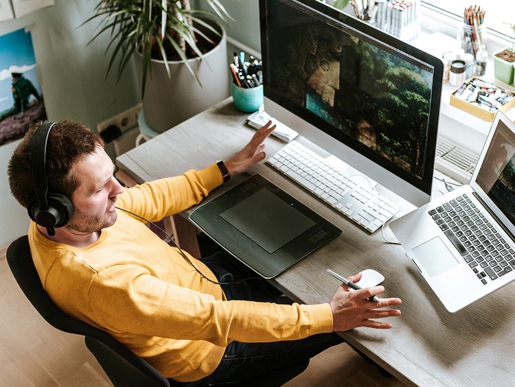 Un hombre con un suéter amarillo y auriculares trabaja en un escritorio con un monitor de escritorio y una laptop.