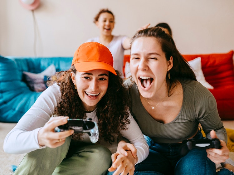 Dos mujeres se ríen entusiasmadas mientras sostienen mandos de videojuegos en un sofá.