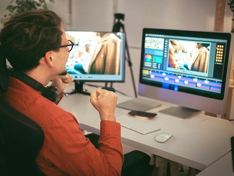 Un hombre con una camisa naranja trabaja en una computadora de escritorio que muestra un software de edición de video.
