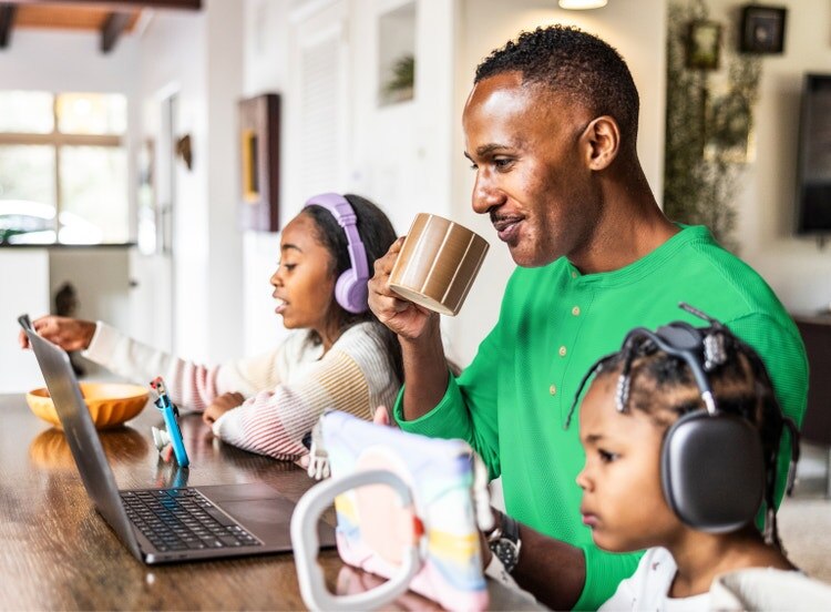 Un padre bebe de una taza en una mesa mientras dos niños usan auriculares y utilizan una laptop y una tableta a su lado.