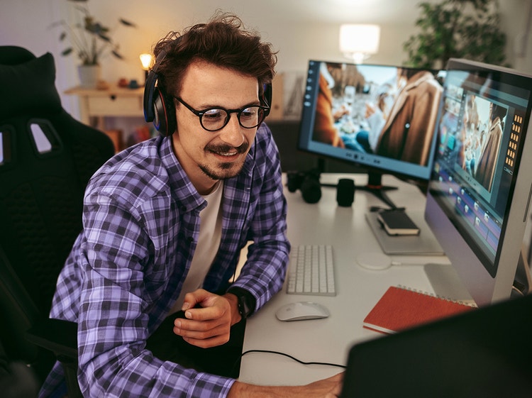 Un hombre con lentes y auricualres trabaja en un escritorio con varias pantallas de computadora.