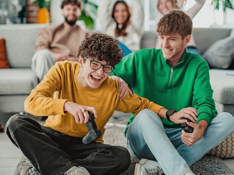 Dos jóvenes están sentados en un sofá, riéndose mientras juegan juntos un videojuego.