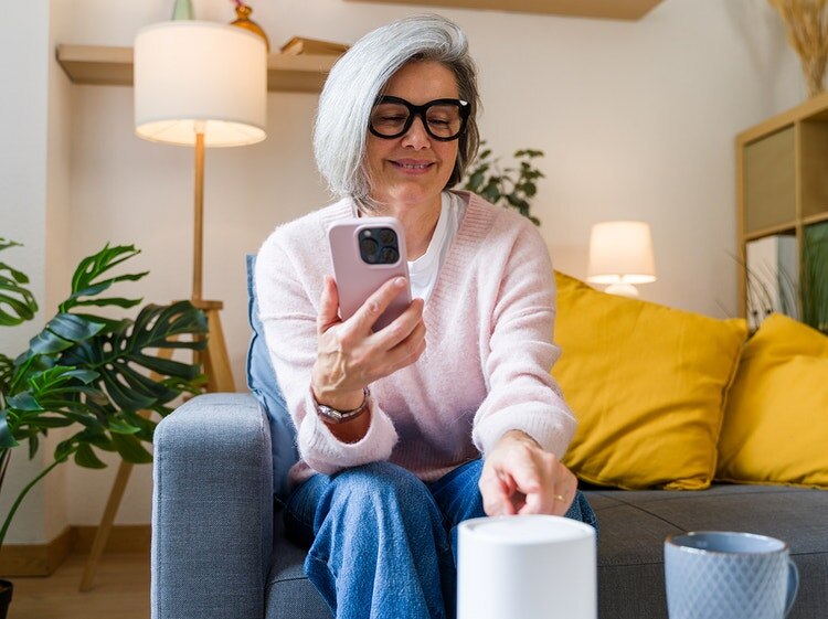 Una mujer mayor está sentada en un sofá y mira su teléfono, mientras que cerca de ella hay un dispositivo de smart home.