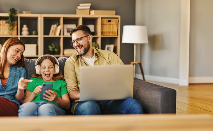 Una familia de tres personas, todas sonrientes, está sentada en un sofá; una niña con auriculares se ríe mirando su teléfono, mientras sus padres la observan.
