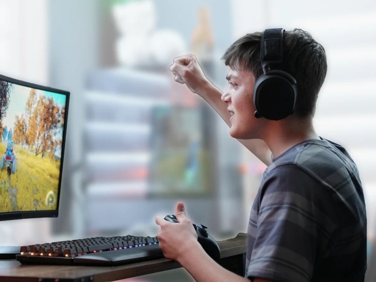 Teenage boy wearing gaming headphones celebrating with a raised fist while playing a video game on a curved monitor, with a controller in his other hand.