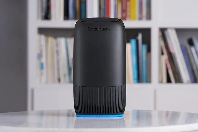 Spectrum Advanced WiFi mesh router on a table with a blue light ring and blurred shelves behind it.