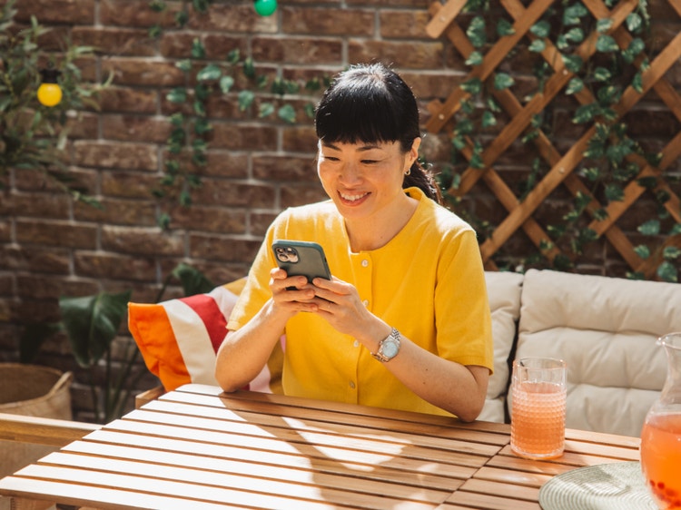 Smiling woman in a yellow shirt using her smartphone while seated at an outdoor table.
