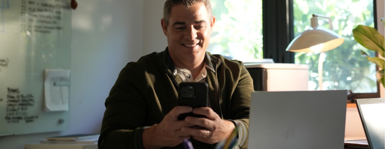 Hombre sonriente con un teléfono móvil y una laptop mientras trabaja desde casa