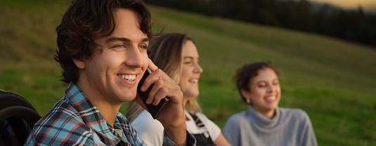 Man with curly hair smiling while on a phone call outdoors, with two women in the background