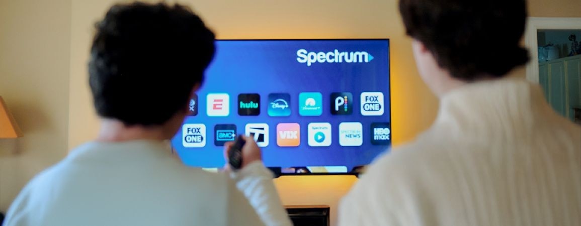A hand pressing a button on a TV remote in front of a TV.