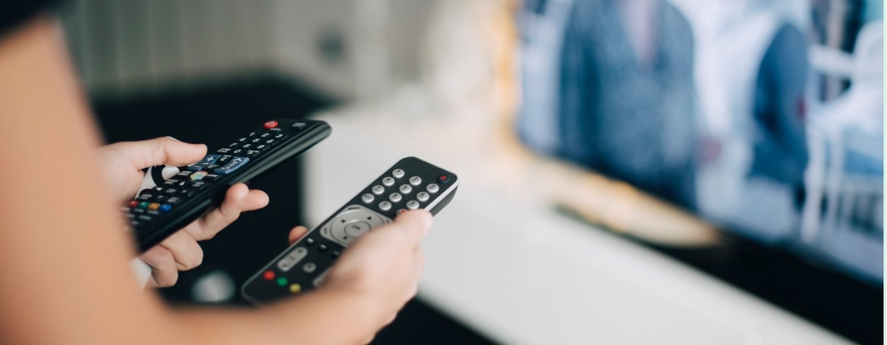 Person holding two TV remote controls with a blurred television screen in the background