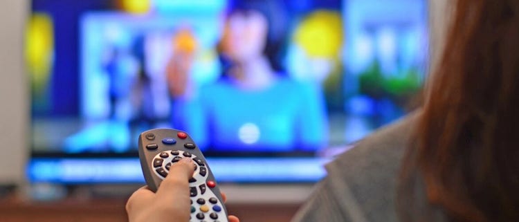 A hand pressing a button on a TV remote in front of a TV.
