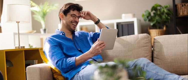 Man in a blue shirt smiling while streaming content on a tablet on his living room couch.