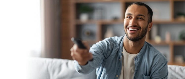 A man sitting on a couch and holding a remote while smiling.