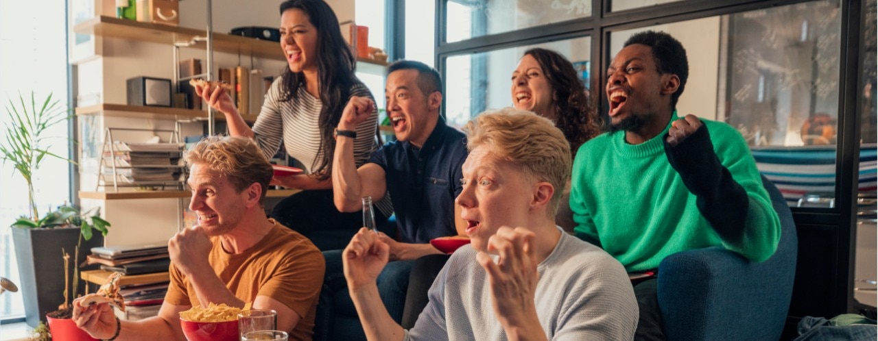 Six friends cheering and eating snacks while watching a sports game. 